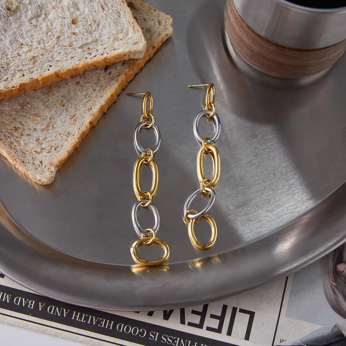 Gold and silver chain earrings on a metallic surface with toast and a cup.